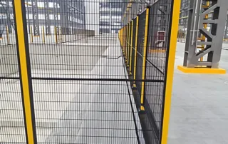 Detalhe do canto da Cerca de Máquina mostrando poste amarelo e malha preta ao redor de uma célula robótica.