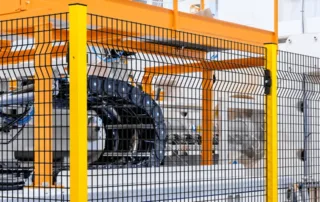 Valla de seguridad sin marco con paneles de malla soldada y postes de acero amarillos protegiendo maquinaria de producción automatizada