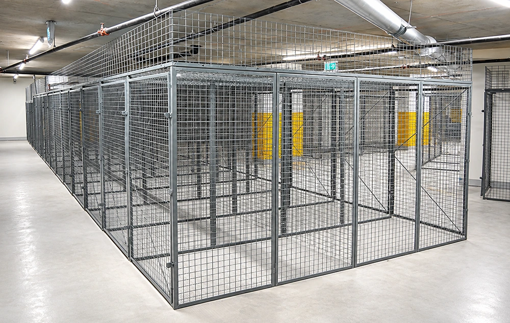 Modular gray steel wire mesh storage cages with hinged padlock doors and cross-braced panels in a warehouse basement